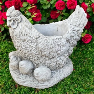 May include: A gray, concrete hen-shaped planter with a textured surface. The planter has a large opening for plants and three egg-shaped feet. The background features red roses and green grass.