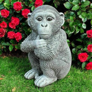 May include: A gray stone monkey statue sitting on green grass, with its hands clasped. Red roses and green foliage are in the background. The monkey has a thoughtful expression.