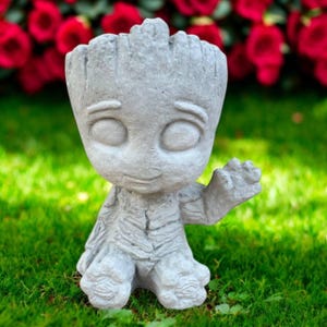 May include: A gray concrete Groot-like planter statue. The statue has a textured bark-like appearance, large oval eyes, and a small smile. It is sitting on green grass with red flowers in the background. The statue is approximately 8 inches tall.