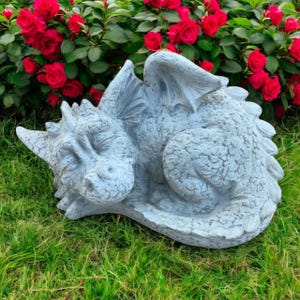 May include: A sleeping dragon statue in a light gray color, with textured details. The dragon has closed eyes and is curled up on green grass, with red roses in the background. A garden ornament.
