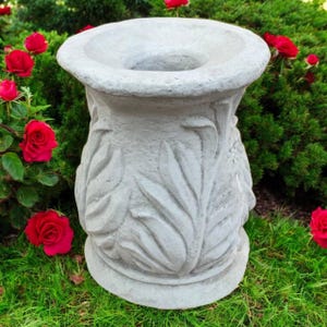 May include: A grey concrete garden planter with a wide, shallow top and a flared base. The planter has a detailed leaf design around the body. Red roses and green foliage are in the background.