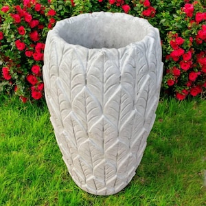 May include: A large, gray concrete planter with a leaf pattern. The outdoor garden pot is oval-shaped and sits on green grass, with red flowers in the background. The planter is designed to hold plants and flowers.