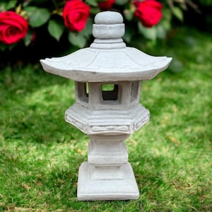 May include: A gray stone Japanese garden lantern with a pagoda-style roof and geometric design. The lantern has a square base and is set on a grassy lawn with red roses in the background.