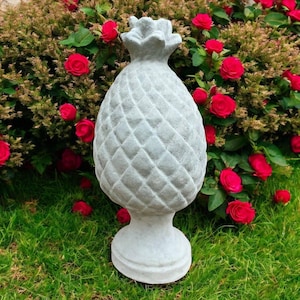 May include: A gray concrete pineapple statue with a textured surface, set on a pedestal base. The statue is surrounded by green grass and a backdrop of red roses and green foliage, creating a garden theme.