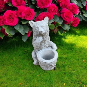 May include: A gray stone-colored bear figurine featuring a textured fur finish and a hollow, cylindrical opening in the front. The bear is depicted in a seated position with one paw raised near its chin, crafted from a solid material.