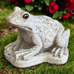 May include: A detailed, gray frog statue with dark eyes sits on a stone base, surrounded by green grass and red roses in the background. The frog sculpture has a textured surface, capturing the details of the frog's skin.
