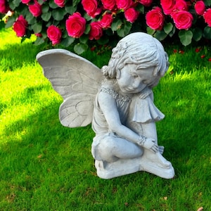 Peut inclure: Statue de fée en béton gris avec de grandes ailes, assise sur de l'herbe verte. La fée regarde vers le bas, les bras autour de ses jambes. Un buisson de roses roses vibrantes est en arrière-plan.