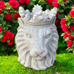May include: A gray concrete lion head sculpture wearing a crown, set against a backdrop of red roses and green grass. The lion's face has detailed features, and the crown adds a regal touch. This garden decor piece is ideal for outdoor spaces.