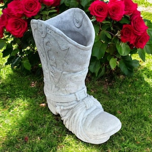 May include: A grey concrete cowboy boot planter with embossed details, including a star and rope accents. The boot is set on green grass with red roses in the background, suitable for outdoor decor.