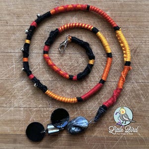 May include: A handmade, colorful hair wrap with a spiral design. The wrap features black, orange, and yellow yarn, accented with small silver bells. A small charm with black circles and a blue stone hangs from the end. The Little Bird logo is in the bottom right corner.
