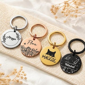 May include: Four circular metal keychains with split rings. The silver one features a floral wreath and the name Maggie. The rose gold one shows flowers and the name Flora. The gold one displays a cat silhouette and Maggie. The black one shows space motifs and Hazel.