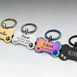 Puede incluir: Cuatro placas de identificación para mascotas en forma de hueso con anillas. La placa dorada dice Simba, la plateada Oscar Call My Family 324-501-4367, la iridiscente Edsel Microchipped y la negra OLIVER. Cada una incluye huellas de patas grabadas.