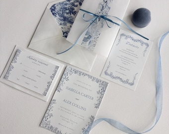 May include: Wedding invitation suite featuring a blue and white floral design. Includes an invitation, RSVP card, details card, and envelope with a matching patterned liner. A blue ribbon and a small blue velvet sphere complete the set.