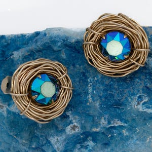 May include: A pair of gold-toned wire nest earrings, each with a faceted blue and green crystal center. The earrings are displayed on a textured, blue stone surface, creating a striking contrast.