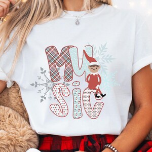 May include: White t-shirt with the word "MUSIC" in a festive design. The letters are in red, blue, and plaid patterns, with a snowflake and an elf graphic. The shirt is a casual, short-sleeved style.