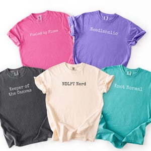 May include: Five colorful t-shirts in pink, purple, teal, beige, and charcoal gray. Each shirt has a different white text design: "Fueled by Floss", "Needleholic", "Keeper of the Canvas", "NDLPT Nerd", and "Knot Normal".