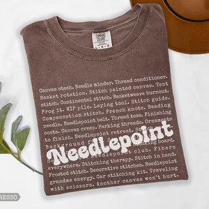 May include: Espresso-colored t-shirt with the word "Needlepoint" in a white, retro font. The shirt features a list of needlepoint-related terms in white text. A brown hat is visible in the upper right corner.