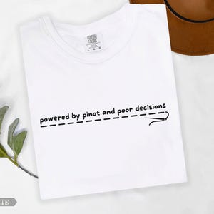 May include: White t-shirt featuring the phrase "powered by pinot and poor decisions" in black, with a dashed line and needle design. A brown hat is partially visible. The t-shirt is a Comfort Colors brand.