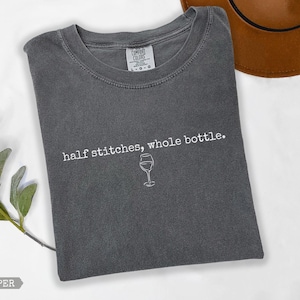 May include: A dark grey t-shirt featuring the text "half stitches, whole bottle." and a wine glass illustration. The shirt is made of a soft material and is a comfortable fit. The shirt is a size L-G.