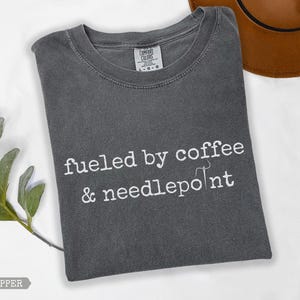 May include: A dark grey t-shirt with the text "fueled by coffee & needlepoint" in white. The shirt is a Comfort Colors brand and has a crew neck. A brown hat is visible in the background.