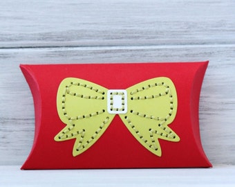 May include: A red gift box with a yellow, hand-embroidered bow design. The box is pillow-shaped and features a white buckle detail. Text on the image reads "Christmas Ribbon Hand-Embroidered Gift Box."