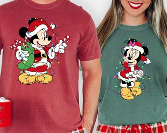 Puede incluir: Dos personas con pijamas navideños a juego. Uno lleva una camiseta granate con un dibujo de Mickey Mouse, el otro una camiseta verde con un dibujo de Minnie Mouse. Ambas camisetas presentan luces festivas y gorros de Papá Noel. Llevan pantalones de pijama de cuadros rojos y verdes.