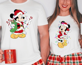 Puede incluir: Camisetas blancas con un diseño de Mickey Mouse con temática navideña. El Mickey de dibujos animados viste un traje de Papá Noel y está rodeado de luces navideñas. Las camisetas se combinan con pantalones de pijama de cuadros rojos. Una persona sostiene una taza roja.