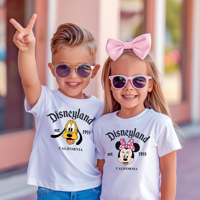 Puede incluir: Dos ni&ntilde;os con camisetas blancas de Disneyland y gafas de sol. El ni&ntilde;o lleva gafas de sol con montura dorada y la camiseta presenta a Pluto. La ni&ntilde;a lleva gafas de sol rosas y un lazo rosa, con un gr&aacute;fico de Minnie Mouse en su camiseta. Ambas camisetas dicen "Disneyland est. 1955 California."