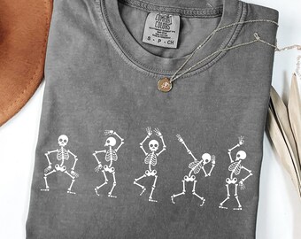May include: A gray Comfort Colors t-shirt featuring a white graphic of five dancing skeletons. The shirt has a crew neck and a small tag. A silver necklace with a pendant is draped across the shirt.