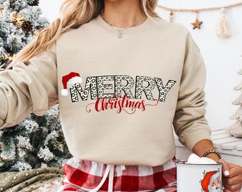 May include: Beige sweatshirt with the text "MERRY Christmas" in a mix of leopard print and red script. A Santa hat graphic is on the left. The person is wearing red and white plaid pajama pants and holding a mug with a Santa Claus graphic.