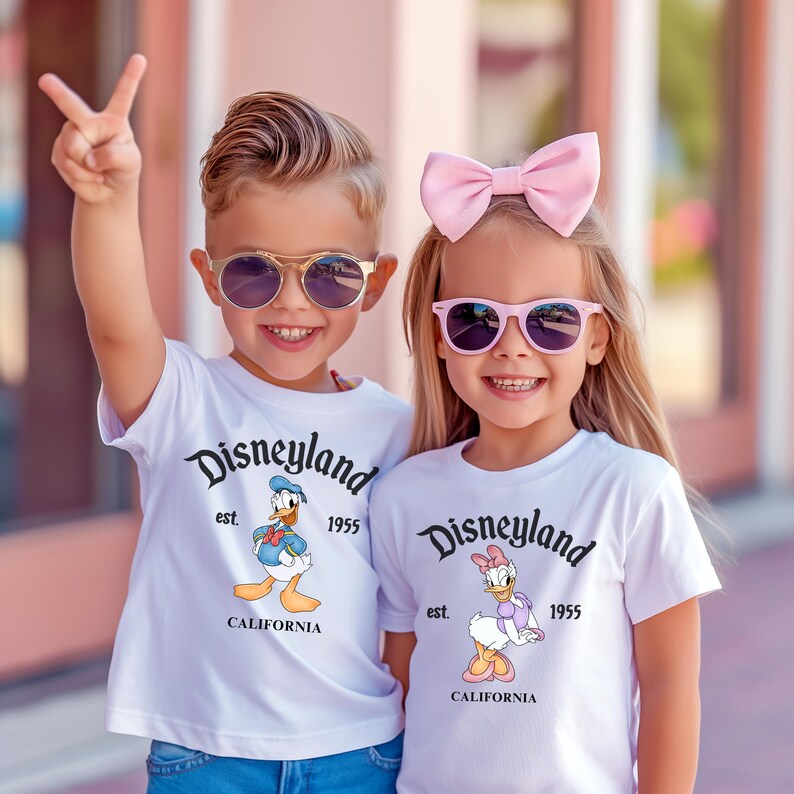 Puede incluir: Dos ni&ntilde;os con camisetas blancas con el texto "Disneyland" y gr&aacute;ficos de personajes de dibujos animados. El ni&ntilde;o lleva gafas de sol y hace el signo de la paz. La ni&ntilde;a lleva un lazo rosa y gafas de sol. Las camisetas tambi&eacute;n dicen "est. 1955" y "California."