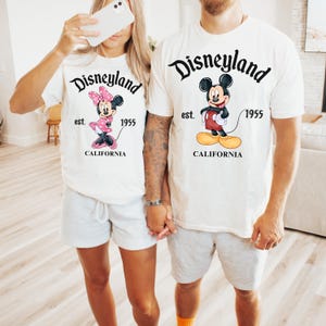 Puede incluir: Dos personas con camisetas blancas a juego con gr&aacute;ficos de Disneyland. Una camiseta presenta a Minnie Mouse en rosa, la otra a Mickey Mouse en rojo. Ambas camisetas tienen impreso "Disneyland est. 1955 CALIFORNIA". Llevan pantalones cortos grises.