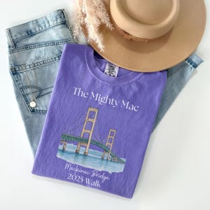 May include: A purple t-shirt with the text "The Mighty Mac" and an illustration of the Mackinac Bridge. The shirt is laid on top of blue jeans, with a tan hat and a gold necklace.