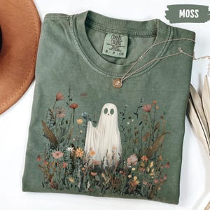 Puede incluir: Una camiseta Comfort Colors verde musgo con un diseño de fantasma en un campo de flores coloridas. La camiseta tiene cuello redondo y mangas cortas. Un collar plateado con un pequeño colgante está drapeado sobre la camiseta.