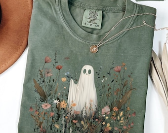 May include: A moss green Comfort Colors t-shirt featuring a ghost design in a field of colorful flowers. The shirt has a crew neck and short sleeves. A silver necklace with a small pendant is draped across the shirt.