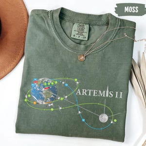 May include: Moss green t-shirt with a graphic of Earth, the moon, and a flight path labeled "Artemis II." The shirt has a crew neck and short sleeves. A gold necklace with a pendant is draped across the shirt.