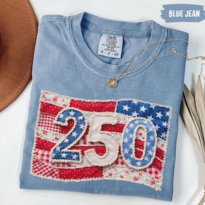 Op de afbeelding: Een blauw Comfort Colors T-shirt met een patriottisch vlagontwerp. De vlag is gemaakt van rood, wit en blauw stof met het nummer 250 in het midden. Een gouden ketting is over het shirt gedrapeerd.