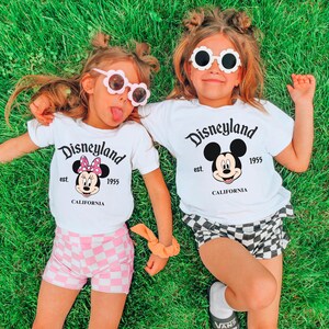 Puede incluir: Dos ni&ntilde;os peque&ntilde;os con camisetas blancas con un gr&aacute;fico de Disneyland y gafas de sol. Uno lleva pantalones cortos de cuadros rosas, el otro pantalones cortos de cuadros blancos y negros. Est&aacute;n tumbados sobre hierba verde.