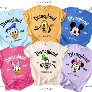 Puede incluir: Una colecci&oacute;n de camisetas coloridas con gr&aacute;ficos de Disneyland. Cada camiseta muestra un personaje diferente de Disney, incluyendo a Pato Donald, Pluto, Mickey Mouse, Daisy y Minnie Mouse. Las camisetas son de varios colores, como azul, amarillo, rosa, blanco y morado.