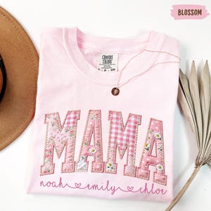 May include: A light pink Comfort Colors t-shirt with the word "MAMA" appliqued in floral and gingham patterns. Below the word "MAMA" are the names "noah", "emily", and "chloe". A gold necklace with a pendant rests on the shirt.