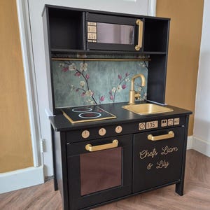 Op de afbeelding: Een zwarte houten speelgoedkeuken met goudkleurige accenten, waaronder handgrepen, een kraan en magnetron details. De keuken heeft een fornuis, een gootsteen, een oven en een magnetron. De tekst "Chefs Liam & Lily" staat op de voorkant.