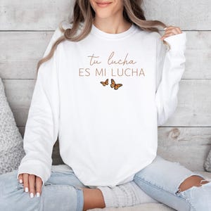 A woman wearing a white long-sleeved shirt with the text "Tú lucha es mi lucha" and butterflies printed on it, sitting on a couch.