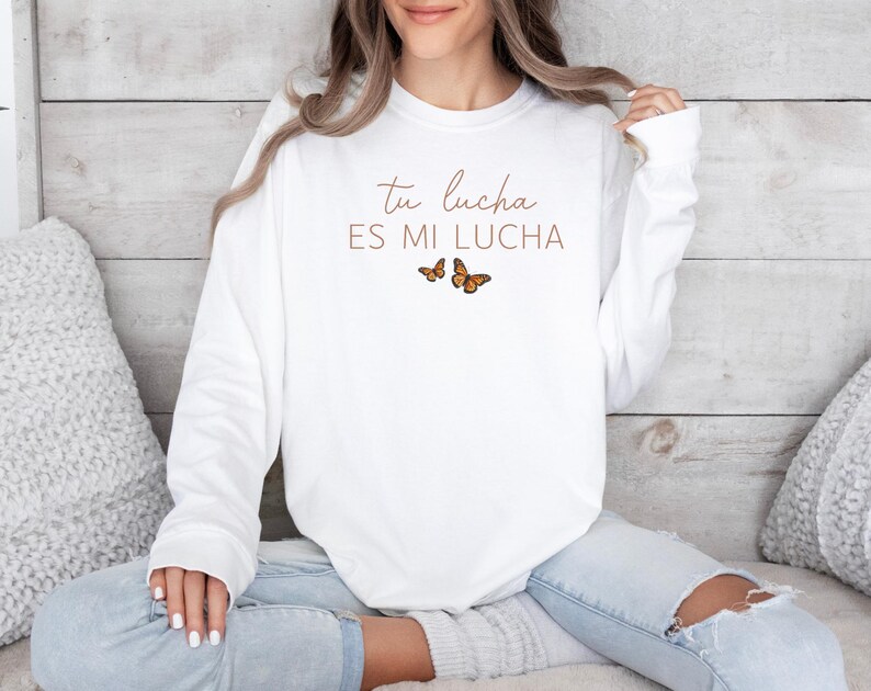 A woman wearing a white long-sleeved shirt with the text "Tú lucha es mi lucha" and butterflies printed on it, sitting on a couch.