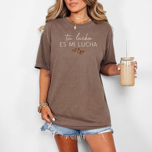 A woman wearing a brown t-shirt with the text "Tú lucha es mi lucha" printed on it, holding a drink in a clear cup.