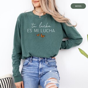 A woman wearing a green long-sleeved shirt with the text "tu lucha es mi lucha" printed on it, standing next to a potted plant.
