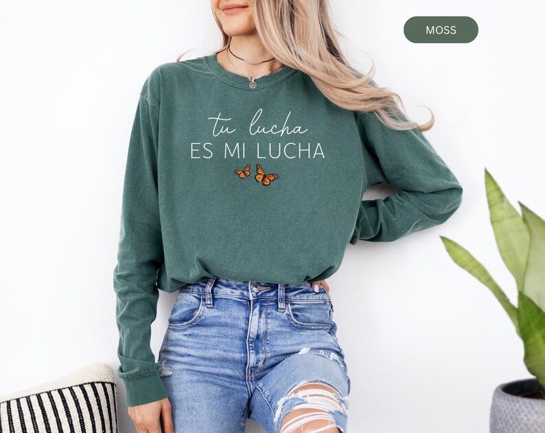 A woman wearing a green long-sleeved shirt with the text "tu lucha es mi lucha" printed on it, standing next to a potted plant.
