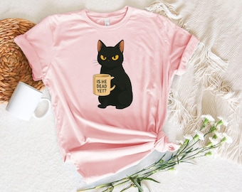 May include: A light pink t-shirt featuring a black cat with yellow eyes holding a mug. The mug has the text "IS HE DEAD YET?". The cat is sitting, and the shirt is knotted at the bottom.