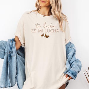 A woman wearing a white t-shirt with the text "tu lucha es mi lucha" and a denim jacket, standing against a white background.