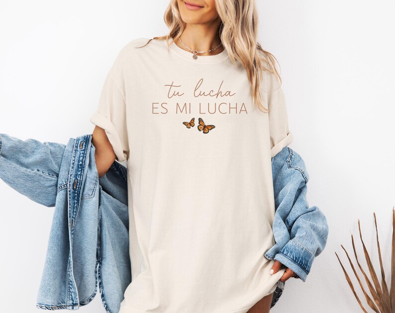 A woman wearing a white t-shirt with the text "tu lucha es mi lucha" and a denim jacket, standing against a white background.