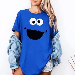 May include: A royal blue t-shirt featuring a large cartoon face with white eyes and a black smile. The shirt is a solid color and has short sleeves. The design is centered on the front of the shirt.